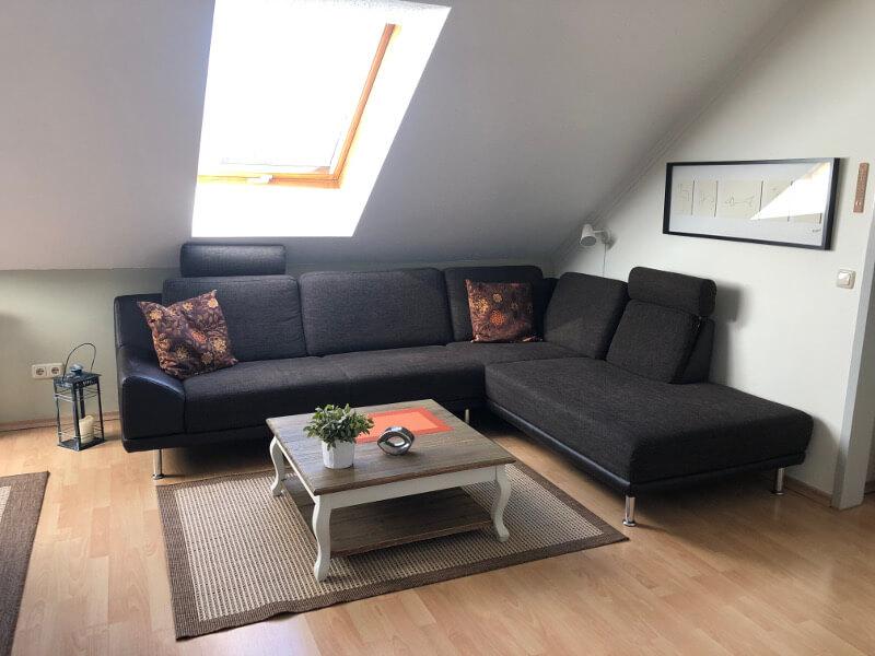A modern living space is presented with a dark sectional sofa adorned with decorative pillows. A light wooden coffee table sits on a woven rug, accompanied by a potted plant. Natural light streams through a skylight, accentuating the room's clean lines and neutral tones.