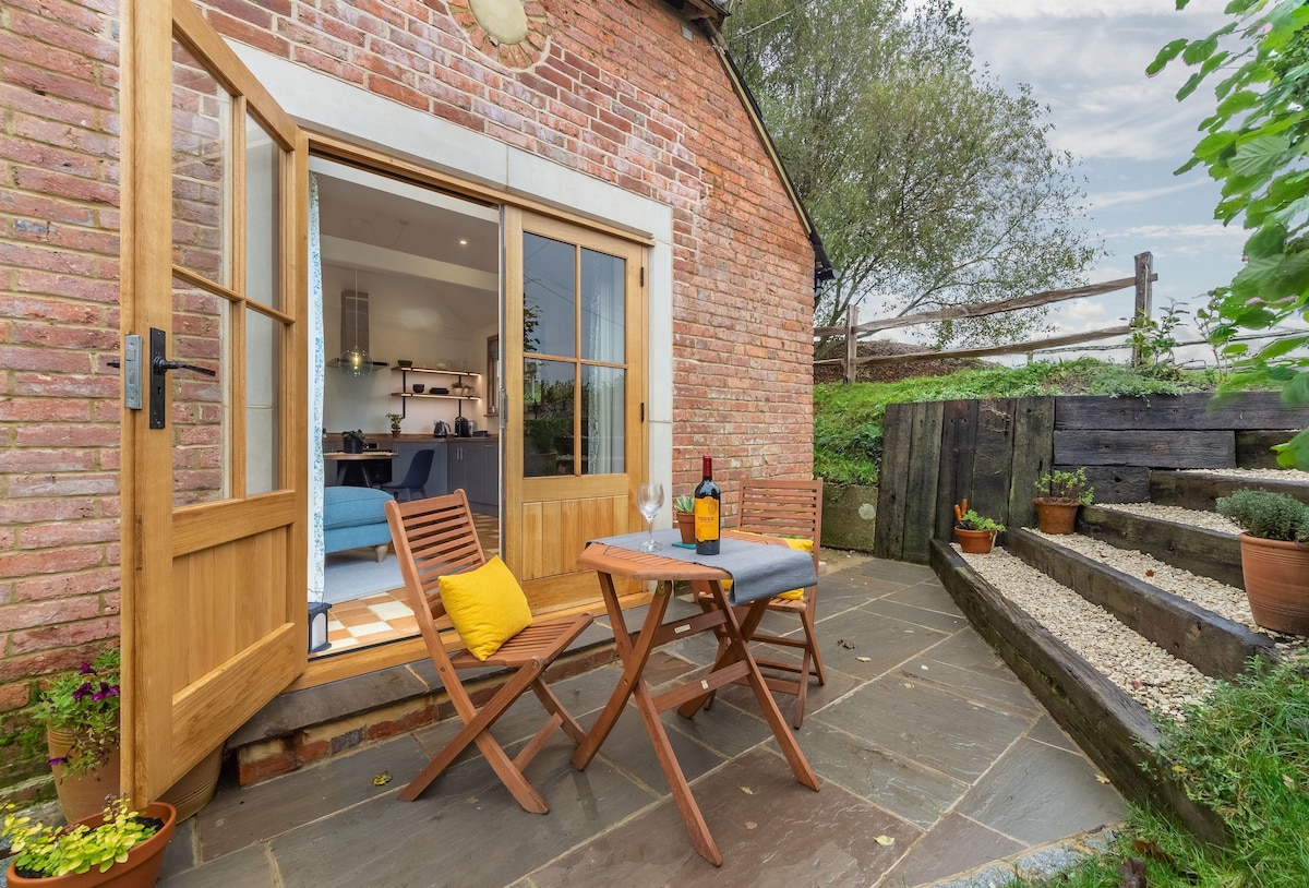 A secluded outdoor patio features two wooden folding chairs and a small table, set against the backdrop of rustic red brick. Potted plants are arranged nearby, and a bottle of wine sits invitingly on the table. The view includes lush greenery and farm fencing.