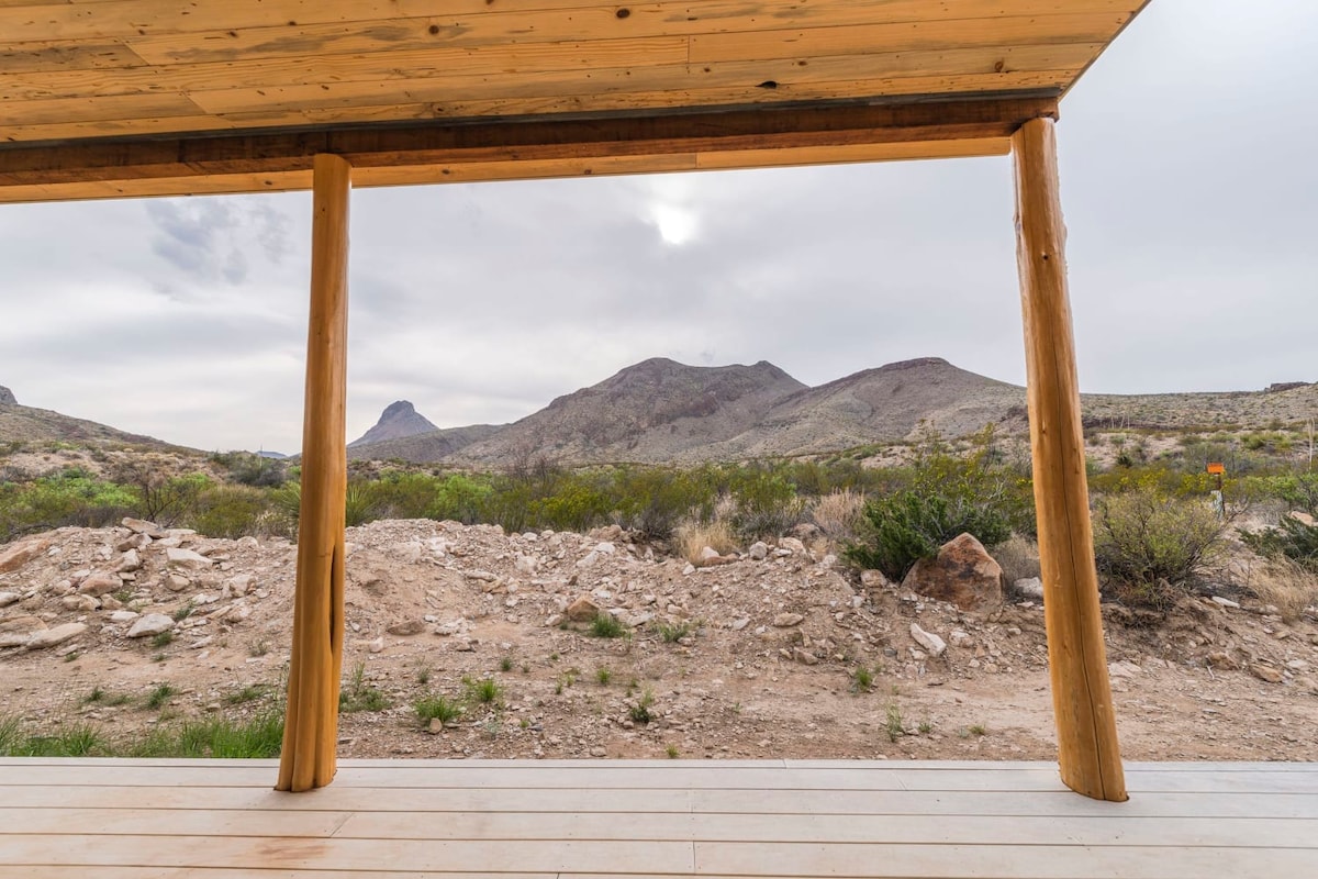 A covered patio is framed by wooden beams, offering an unobstructed view of the rugged mountains and desert landscape. The ground is rocky with sparse vegetation, while a soft overcast sky adds a muted light to the scenery.