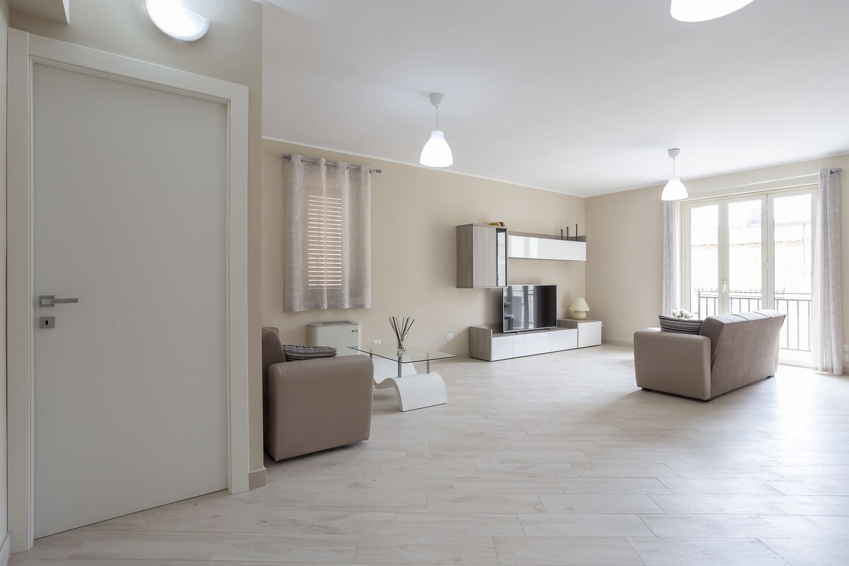 A spacious living area features a light-colored tile floor and neutral walls. Two comfortable couches frame the space, complemented by a modern entertainment unit housing a television. Natural light fills the room through large windows, enhancing the airy ambiance.
