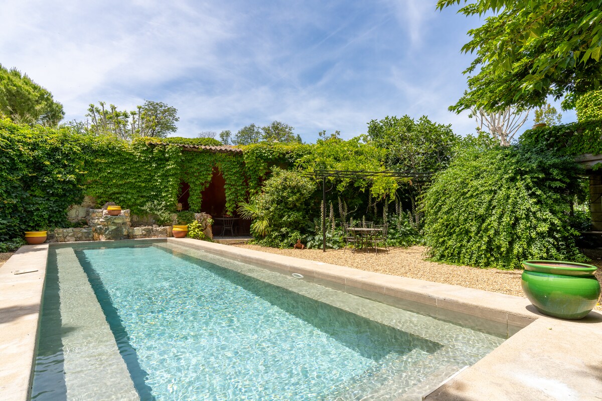 A serene swimming pool is surrounded by lush greenery, featuring stone walls adorned with climbing vines. The clear water reflects the blue sky, while terracotta pots add a touch of color. An inviting seating area is partially visible under a shaded pergola in the background.