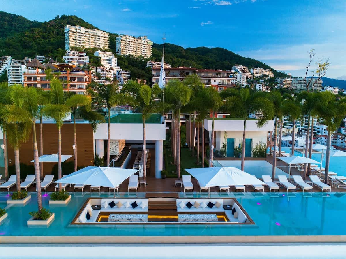 The rooftop pool features an infinity edge, surrounded by lounge chairs and umbrellas. Lush palm trees add greenery, while views extend over the city and hillside. A stylish bar area complements the space, creating an inviting atmosphere for relaxation.