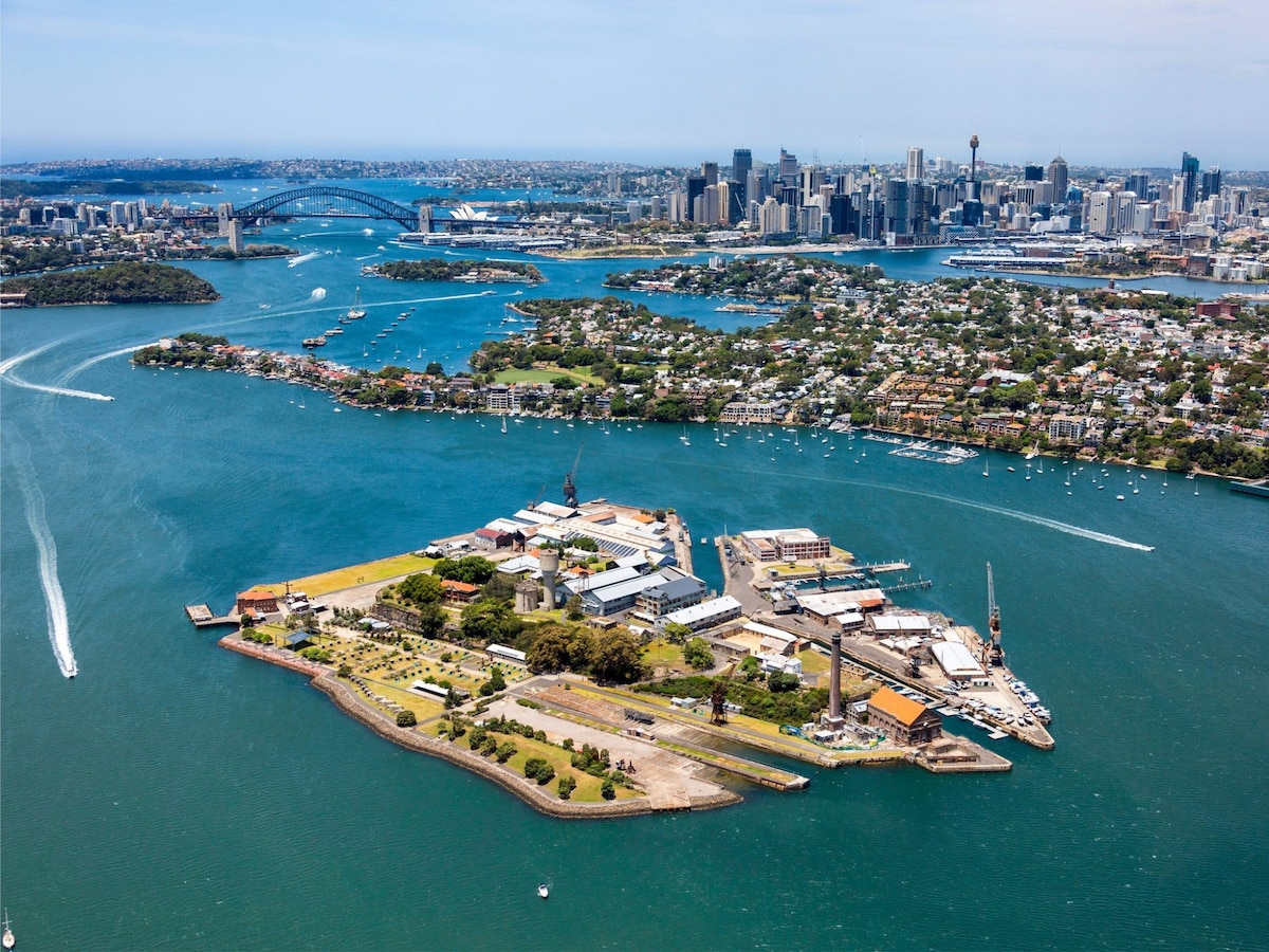 Aerial view of Cockatoo Island, showcasing its distinct shape and surrounding waters. The Sydney skyline and Harbour Bridge are visible in the background, framed by lush greenery and residential areas along the shoreline.