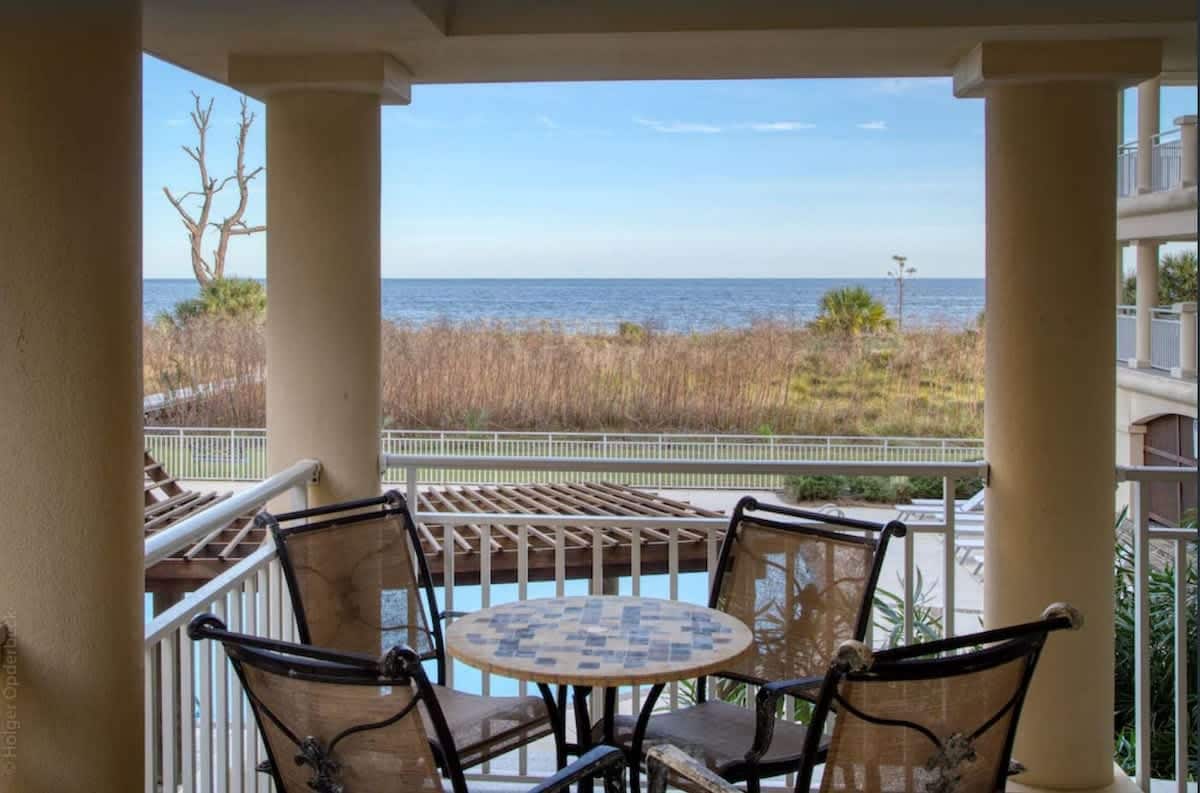 A private balcony features a round table surrounded by metal chairs, providing a serene spot to enjoy views of the ocean. The landscape includes coastal vegetation and a glimpse of the nearby pool area, under a clear blue sky.