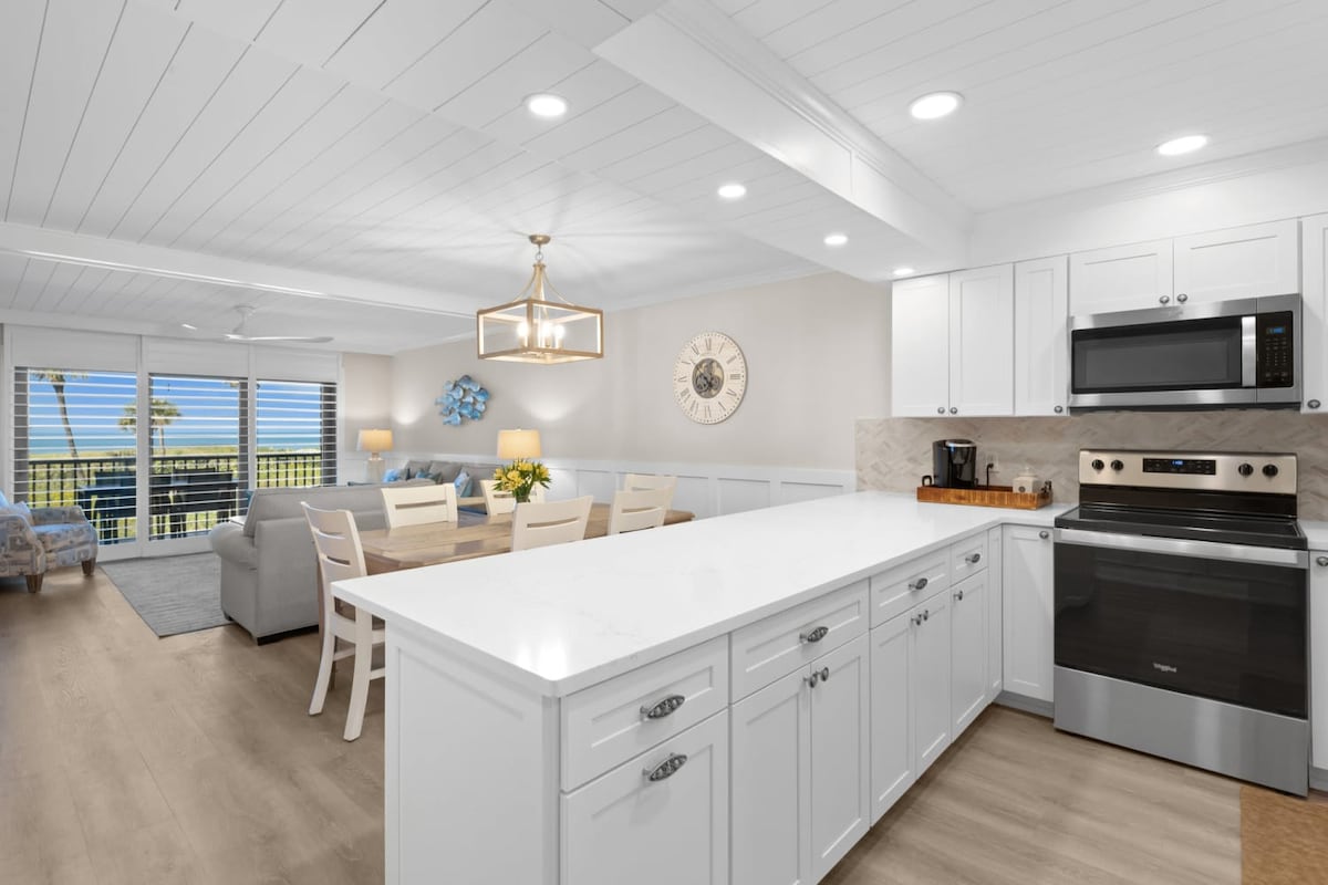 A modern kitchen features sleek white cabinetry and a large island with a light marble countertop. Stainless steel appliances are present, alongside a coffee maker. The open layout connects to a dining area, illuminated by natural light flowing in from large windows showcasing palm trees outside.