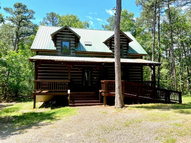 A log cabin features a green metal roof and wooden porches that invite relaxation. Surrounding trees provide a natural backdrop, enhancing the sense of privacy. A gravel path leads to the entrance, while a gentle slope offers accessibility.