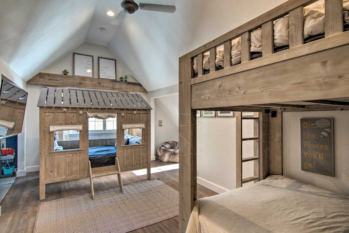 A children's bunk room features a unique wooden playhouse structure with a ladder and sleeping area on the top bunk. A lower bunk with a full mattress provides additional sleeping space. Bright light enters through the windows, enhancing the playful atmosphere.