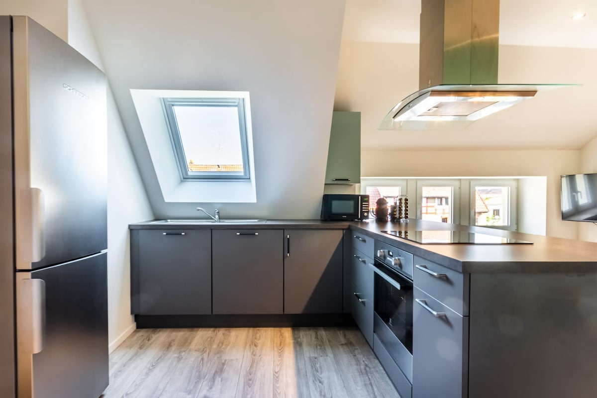 A modern kitchen features a combination of dark cabinetry and stainless-steel appliances, including a refrigerator and an oven. A sloped ceiling enhances the space, while a skylight allows natural light to illuminate the room. An island with a cooktop offers additional preparation space.