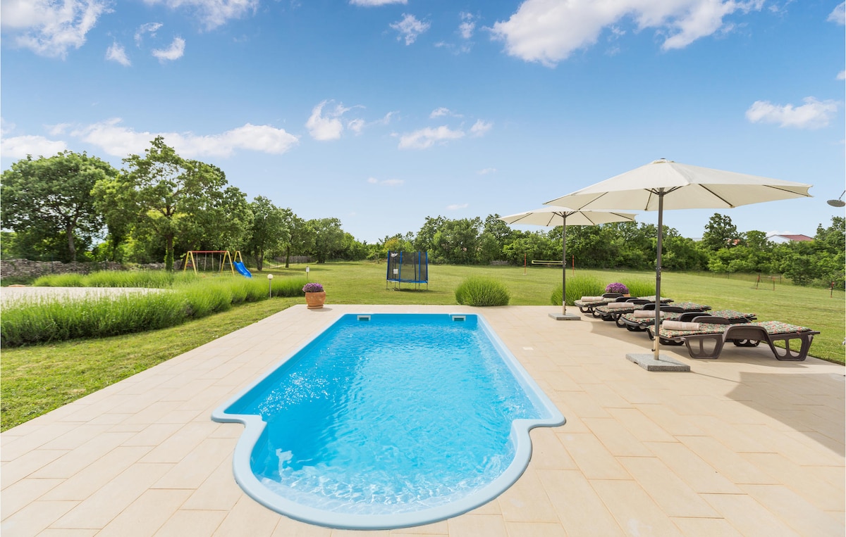 A clear and inviting pool is framed by a spacious patio featuring sun loungers. Shade is provided by large umbrellas, with lush greenery surrounding the area. A playground structure is visible in the background, set against the expansive landscape of the property.