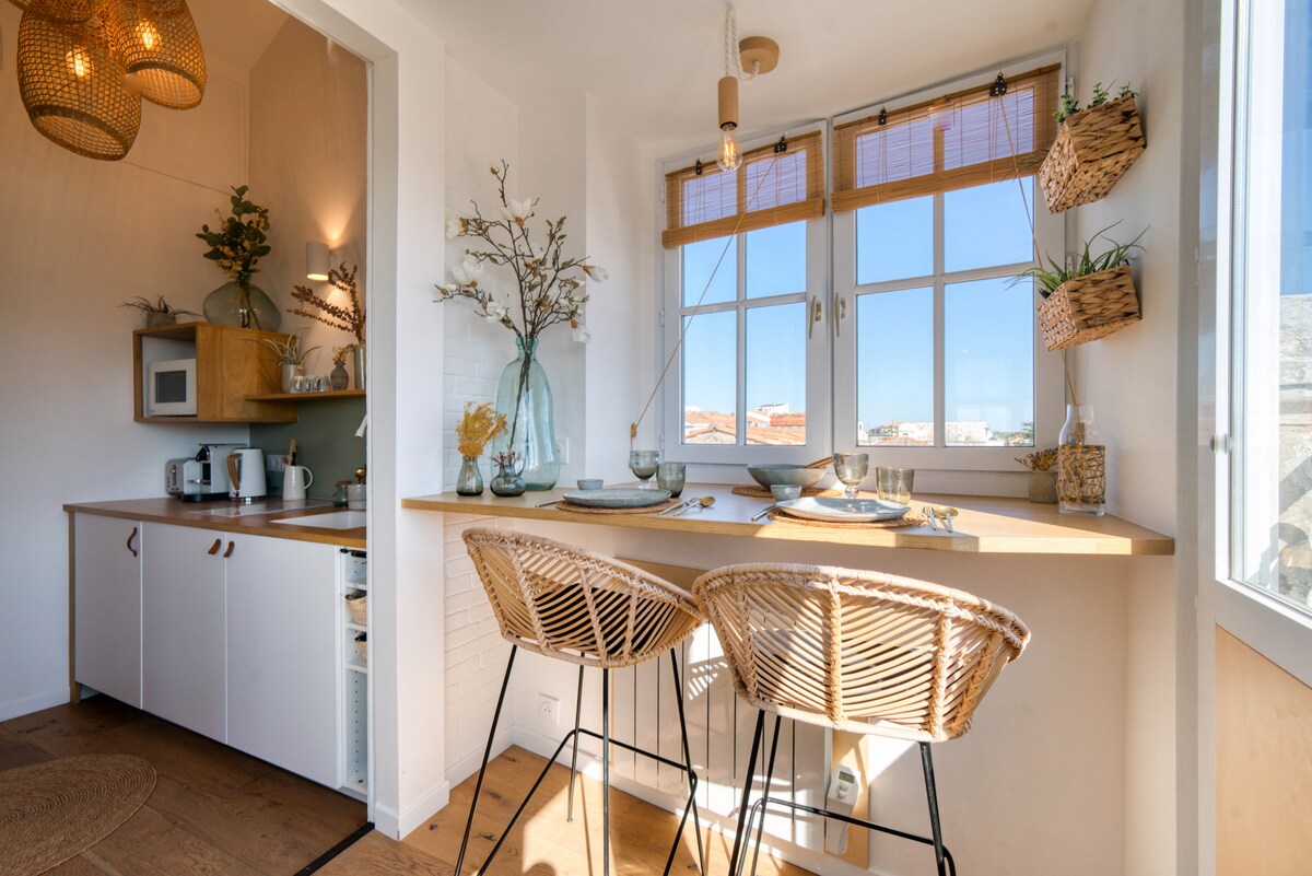 A well-appointed kitchen area features a wooden counter with two woven bar stools. Large windows allow for ample natural light, complementing the decor which includes a decorative vase and small plants. Bright white walls enhance the inviting atmosphere.