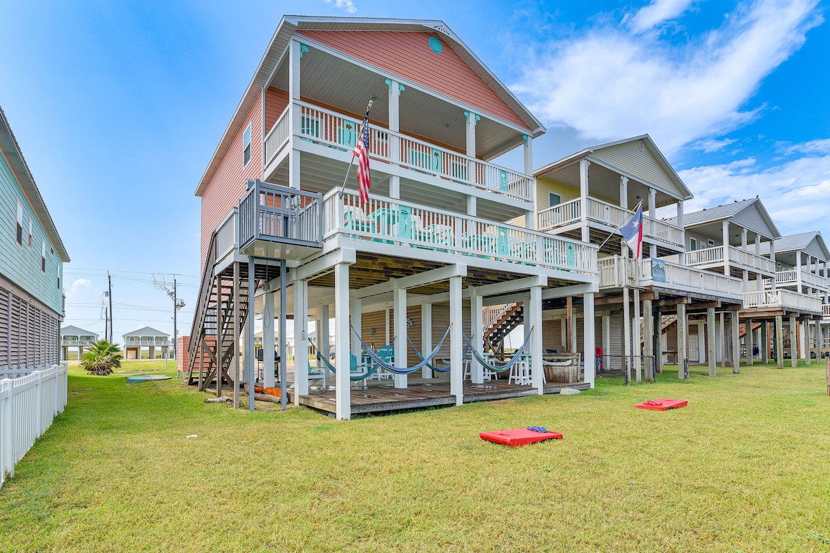 The two-story vacation home is positioned on a grassy area, featuring a spacious deck with multiple lounge areas and hammocks. A staircase leads to the elevated deck, while vibrant flags hang from the porch. Nearby structures are visible in the background against a clear blue sky.