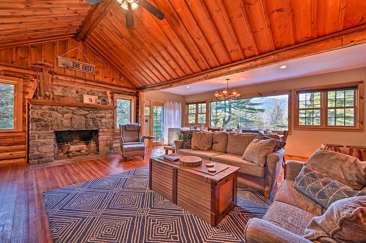 A spacious living area showcases a stone fireplace surrounded by comfortable seating. Natural wood beams highlight the vaulted ceiling. Large windows allow for ample natural light and views of the outdoor landscape. A patterned area rug defines the seating space in front of the coffee table.