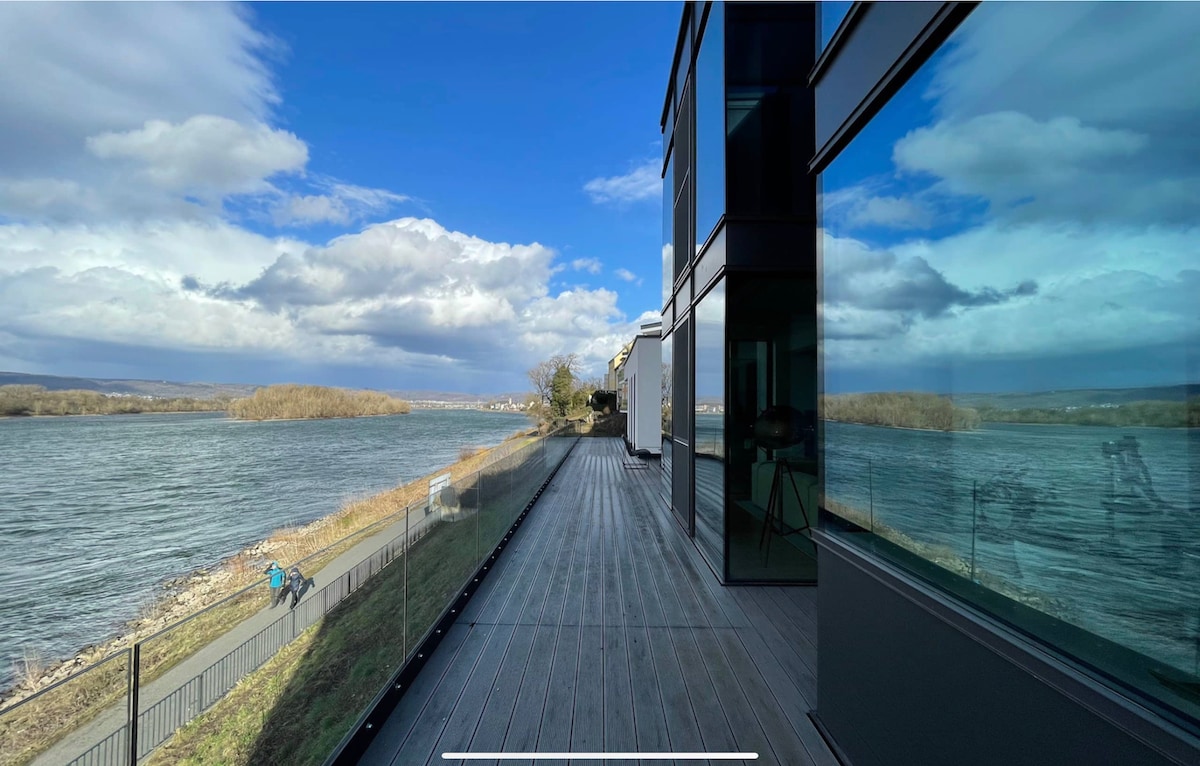 A wide terrace with a wooden deck overlooks the river, reflecting the blue sky and clouds. Large glass doors provide an unobstructed view of the water, creating a seamless connection between indoor and outdoor spaces.