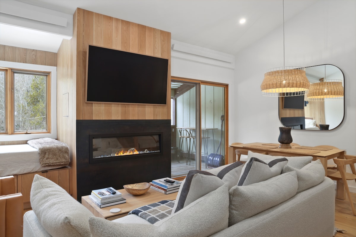 A modern living area features a centrally located electric fireplace, accompanied by a flat-screen television mounted above. Natural light floods in through large windows. A comfortable sofa with neutral tones is positioned around a wooden coffee table, with a dining area illuminated by a woven pendant light.