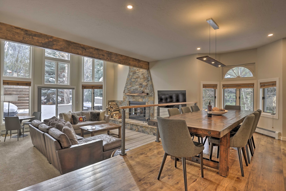 A spacious living area features a stone fireplace as a focal point, surrounded by large windows letting in natural light. A dining table with multiple chairs sits adjacent, creating a welcoming space for meals or conversations. Soft furnishings provide comfort and warmth.