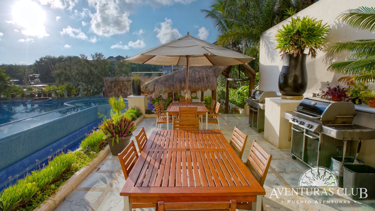 An outdoor dining area is presented with wooden tables and chairs, set beneath a shaded umbrella. A barbecue grill is positioned nearby, surrounded by lush greenery. A swimming pool is visible in the background, complementing the serene atmosphere of the space.