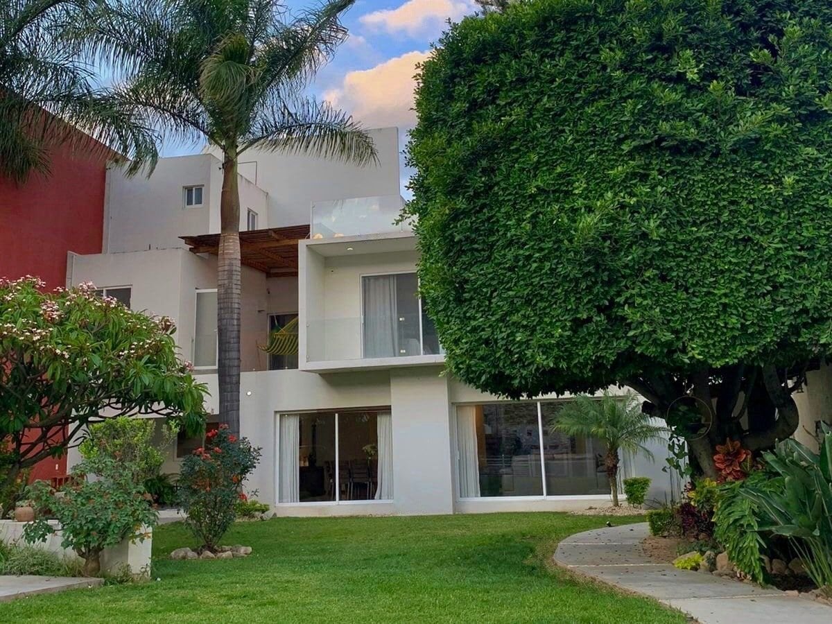 A modern two-story house is framed by a well-maintained garden featuring lush greenery and vibrant flowers. Large glass doors on the ground floor provide views into the spacious interior, while a balcony is visible on the upper level, enhancing the property's inviting design.