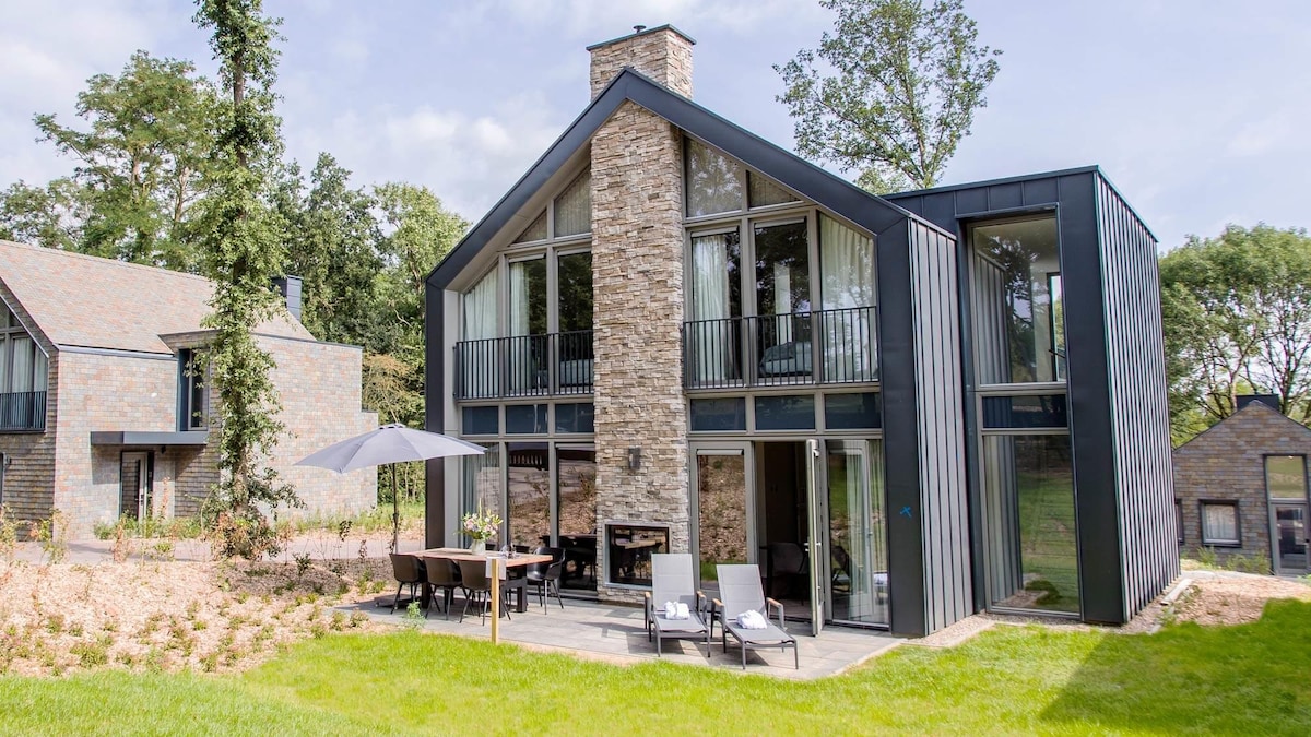 The exterior of a contemporary villa is presented, showcasing a combination of stone and modern materials. Large glass panels create an openness, while a spacious outdoor terrace is furnished with seating. Lush greenery surrounds the property, adding to its serene setting.