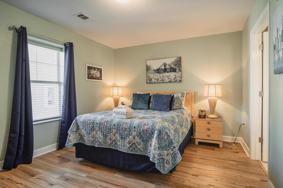 A cozy bedroom features a spacious bed dressed in a patterned blue coverlet, complemented by coordinating pillows. Two bedside lamps illuminate the room, while a large window dressed with dark curtains invites natural light. Artwork hangs above the bed, enhancing the inviting ambiance.