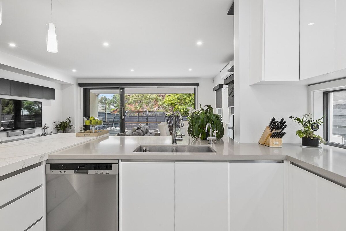 The kitchen features sleek white cabinetry and a large island bench with stone countertops. Stainless steel appliances, including a dishwasher, are visible. Large windows provide natural light and outdoor views, enhancing the open ambiance of the space.