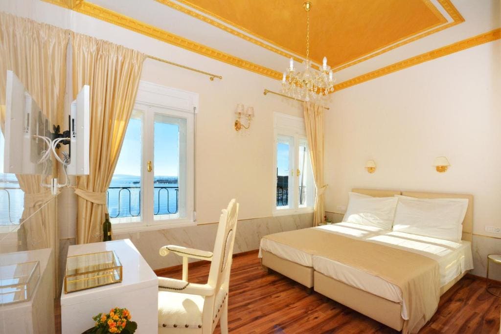 A bright and airy bedroom features large windows offering a view of the sea. A spacious bed with light linens is complemented by a desk and chair, while elegant curtains frame the windows. A chandelier contributes to the room's sophisticated decor.