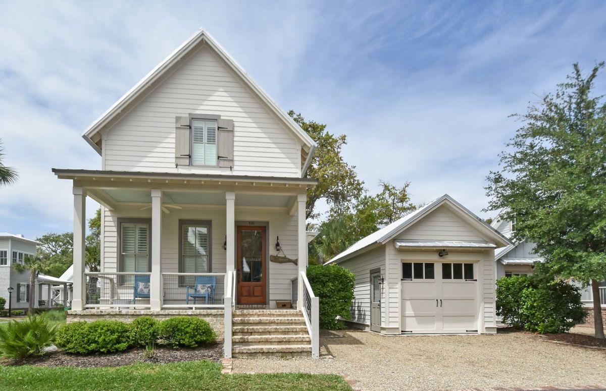 A two-story house is presented with a charming exterior featuring a welcoming front porch. Stone steps lead up to the porch, which is adorned with blue rocking chairs. A detached garage and landscaped area with low-maintenance shrubs are visible, providing a comfortable setting.