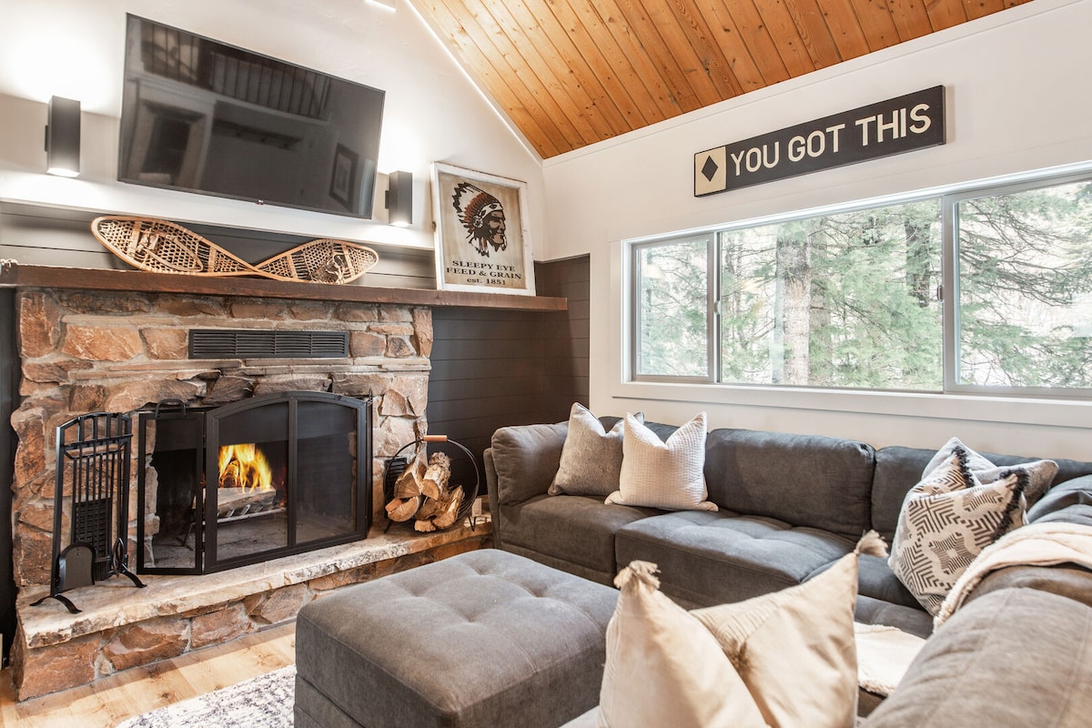 A cozy living room features a stone fireplace with a wood-burning fire, surrounded by a comfortable sectional sofa with multiple throw pillows. A large television is mounted above the fireplace, and natural light streams in through a wide window framed by trees.