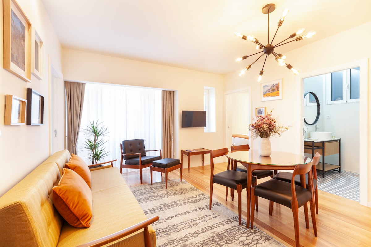 The living area showcases a cozy seating arrangement with a sofa and armchair, complemented by a wooden dining table surrounded by four chairs. Natural light filters through sheer curtains, and a decorative plant adds a touch of greenery. A flat-screen TV is mounted on the wall.