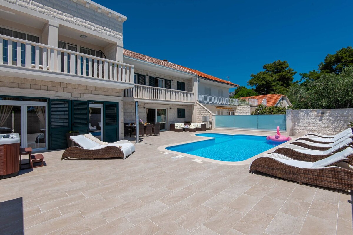 The outdoor area features a private swimming pool surrounded by sun loungers and a patio seating arrangement. A Jacuzzi is positioned to the side. The villa's two-story structure showcases large glass doors leading into the interior, with a backdrop of clear blue skies.