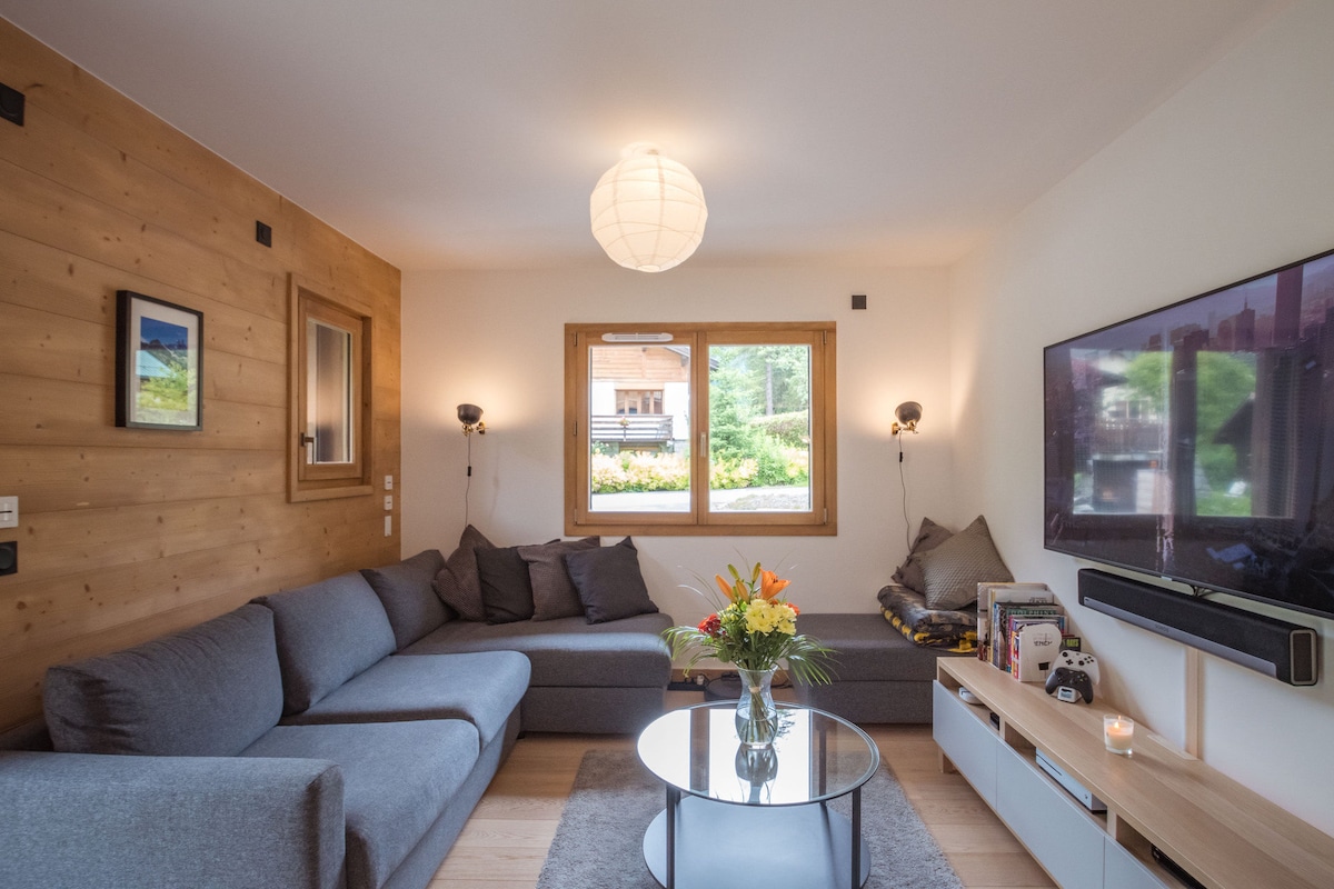 The living area features a spacious gray sectional sofa arranged around a low round table. Soft lighting from wall sconces complements natural light entering through large windows. Decorative items include a floral arrangement on the table and art in the framed images on the wall.