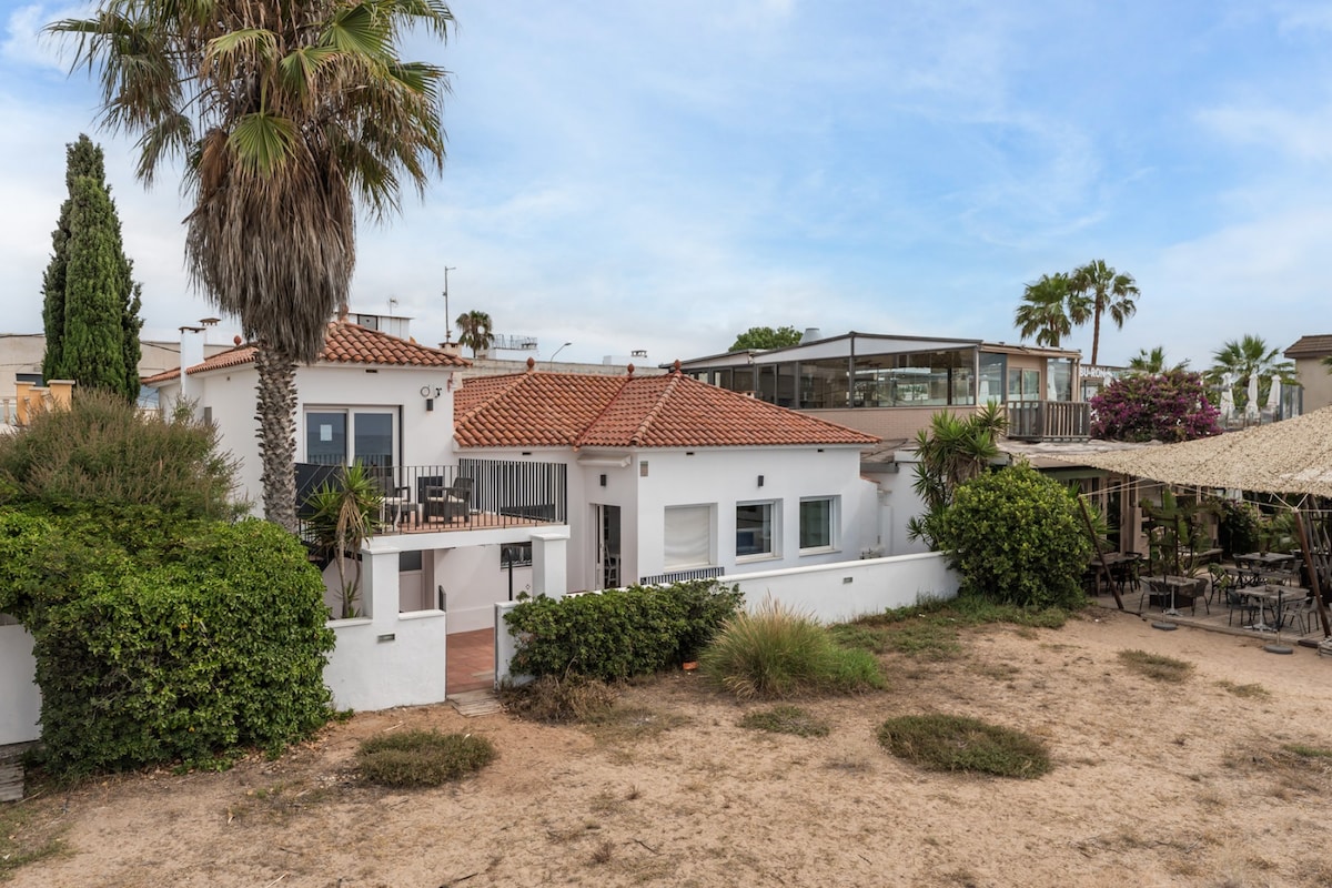 A holiday home is seen with a terracotta tiled roof and surrounded by lush greenery. A private outdoor area with sandy landscaping and a neighboring restaurant can be observed in the background. Tropical plants and palms provide a touch of nature to the setting.