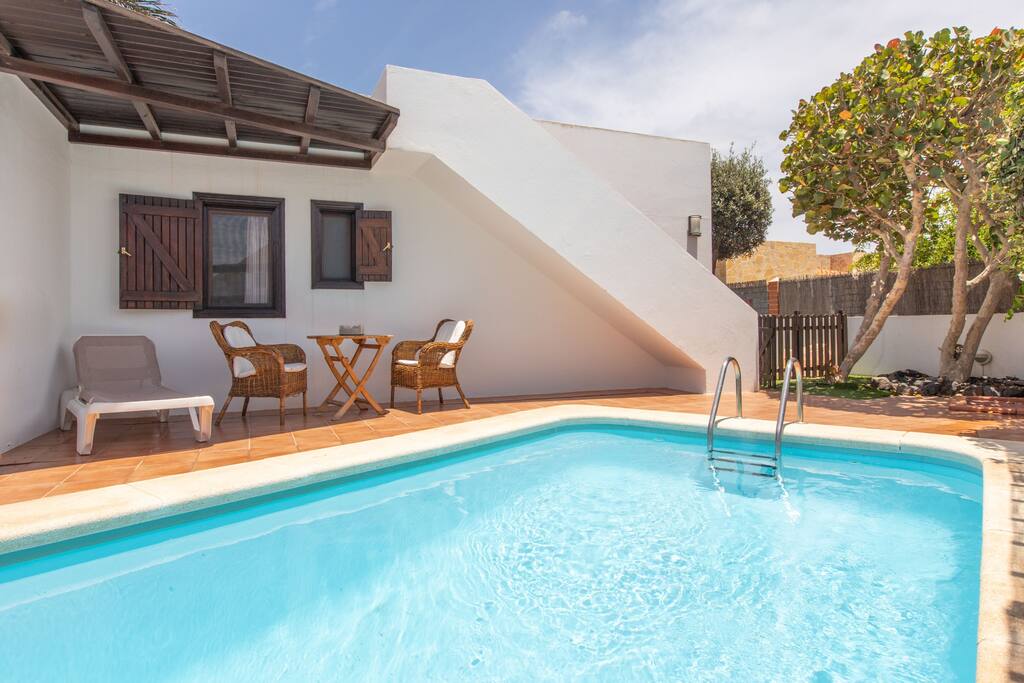 A private heated pool is surrounded by a tiled terrace, featuring two wicker chairs and a small table nearby. The white walls of the villa and a stairway leading to the upper level create a bright and inviting outdoor setting.