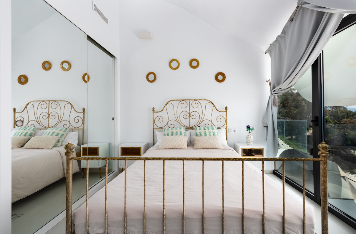 Dormitorio con cama doble y cabecero dorado, ropa de cama, cojines y toallas, mesitas con lámparas, armario con espejos, pared decorada con marcos circulares, alfombra y gran ventanal acristalado con cortinas que conecta con terraza frente al mar.