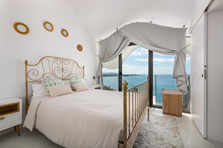 Bedroom with double bed and golden headboard, bed linen, cushions and towels, tables with lamps, wardrobe with mirrors, wall decorated with circular frames, carpet and large glazed window with curtains that connects to the terrace facing the sea.
