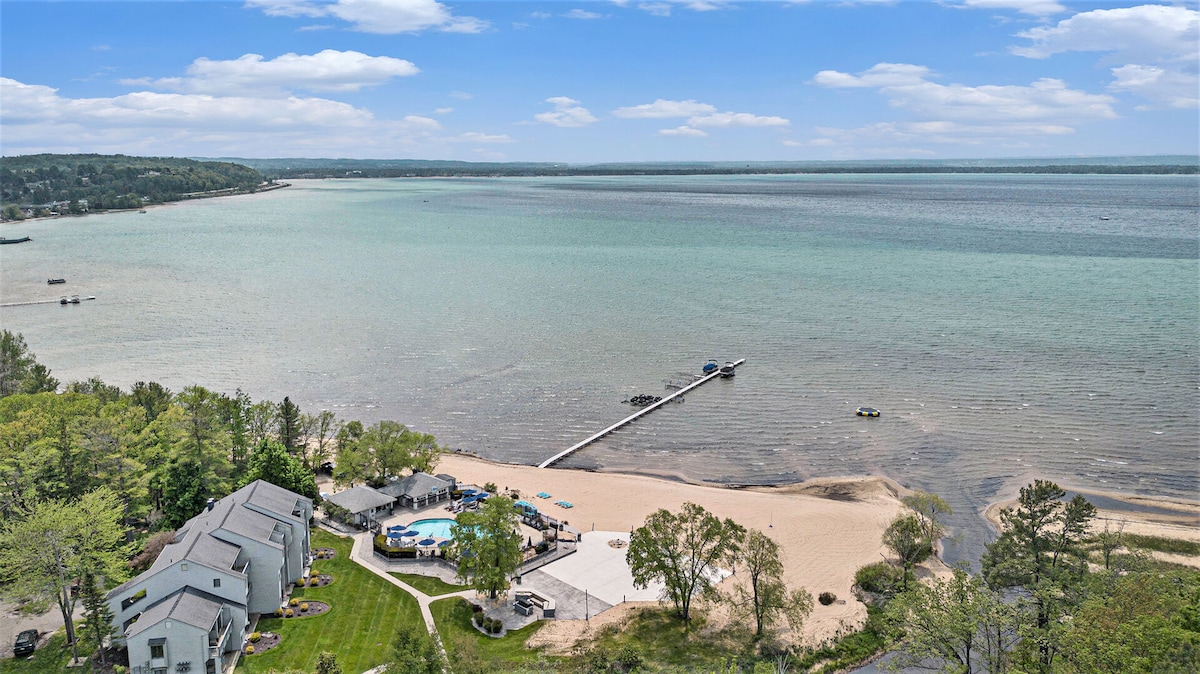 Aerial view of a sandy beach alongside a tranquil body of water, featuring a wooden dock extending into the bay. The shoreline is lined with trees and grassy areas, while several boats can be seen on the water in the distance.