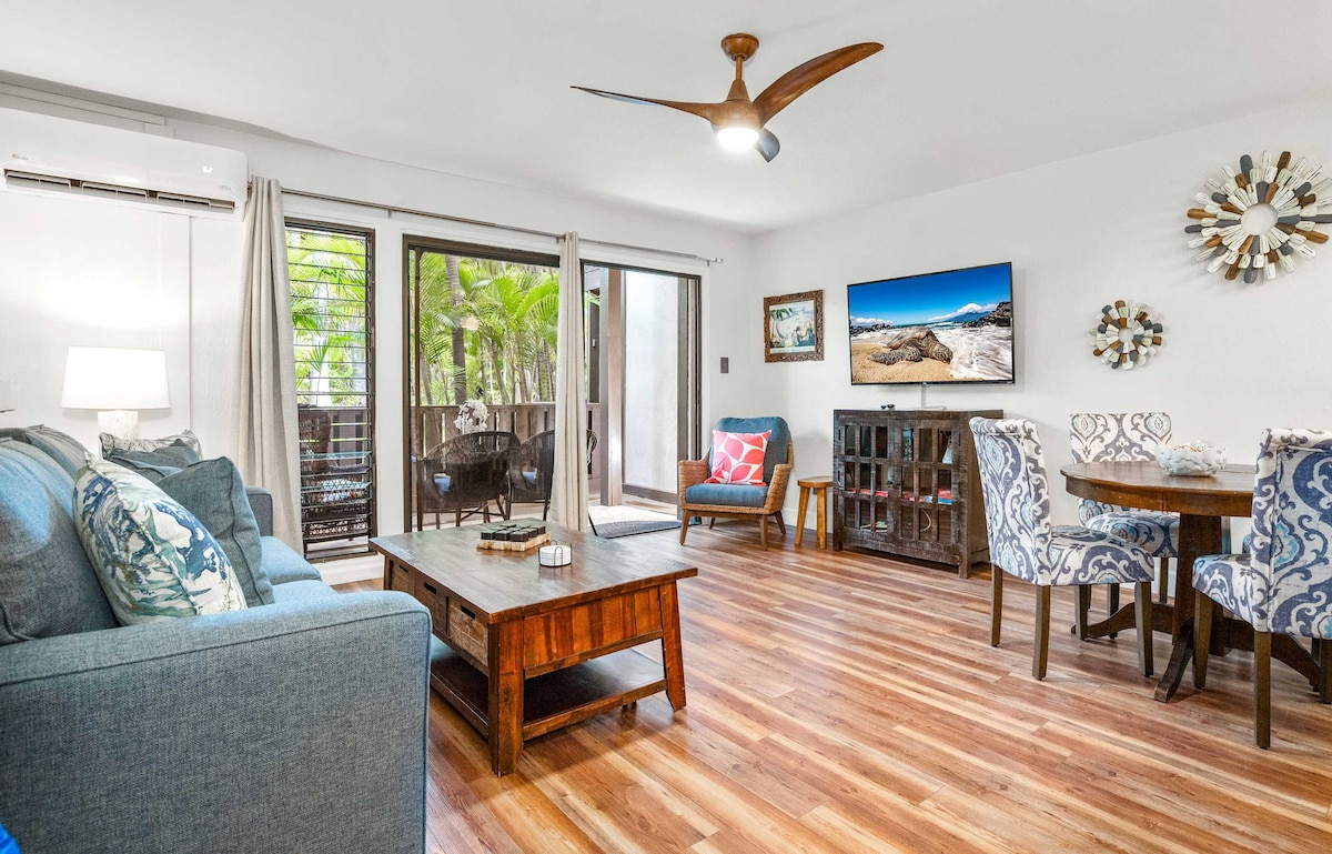 A comfortable living area is showcased with a blue sofa and wooden coffee table. A flat-screen TV is mounted on the wall, and large sliding glass doors open to a patio, allowing natural light in. A round dining table with four upholstered chairs is also present.