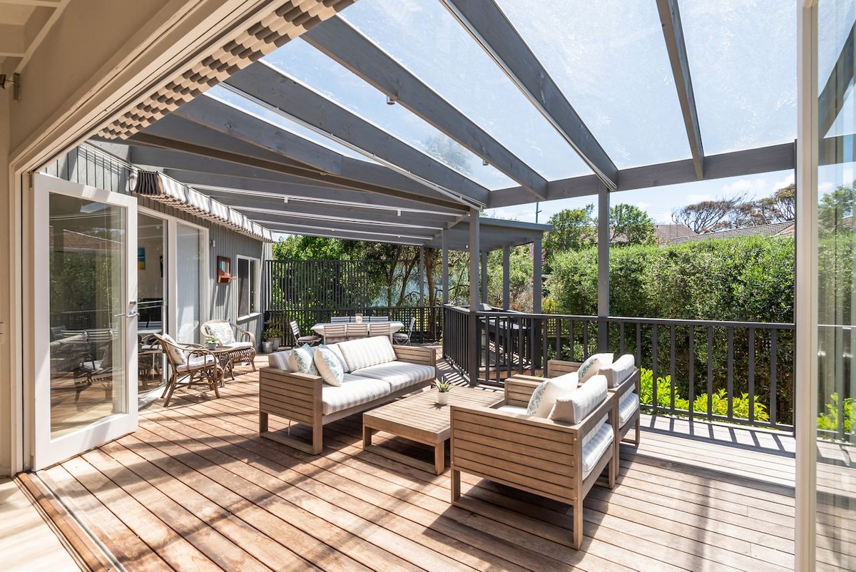 An expansive outdoor deck is featured, offering a spacious seating area with comfortable sofas and armchairs. The area is partially shaded by a transparent roof, providing a bright yet sheltered environment. Lush greenery is visible in the background, enhancing the tranquil atmosphere.
