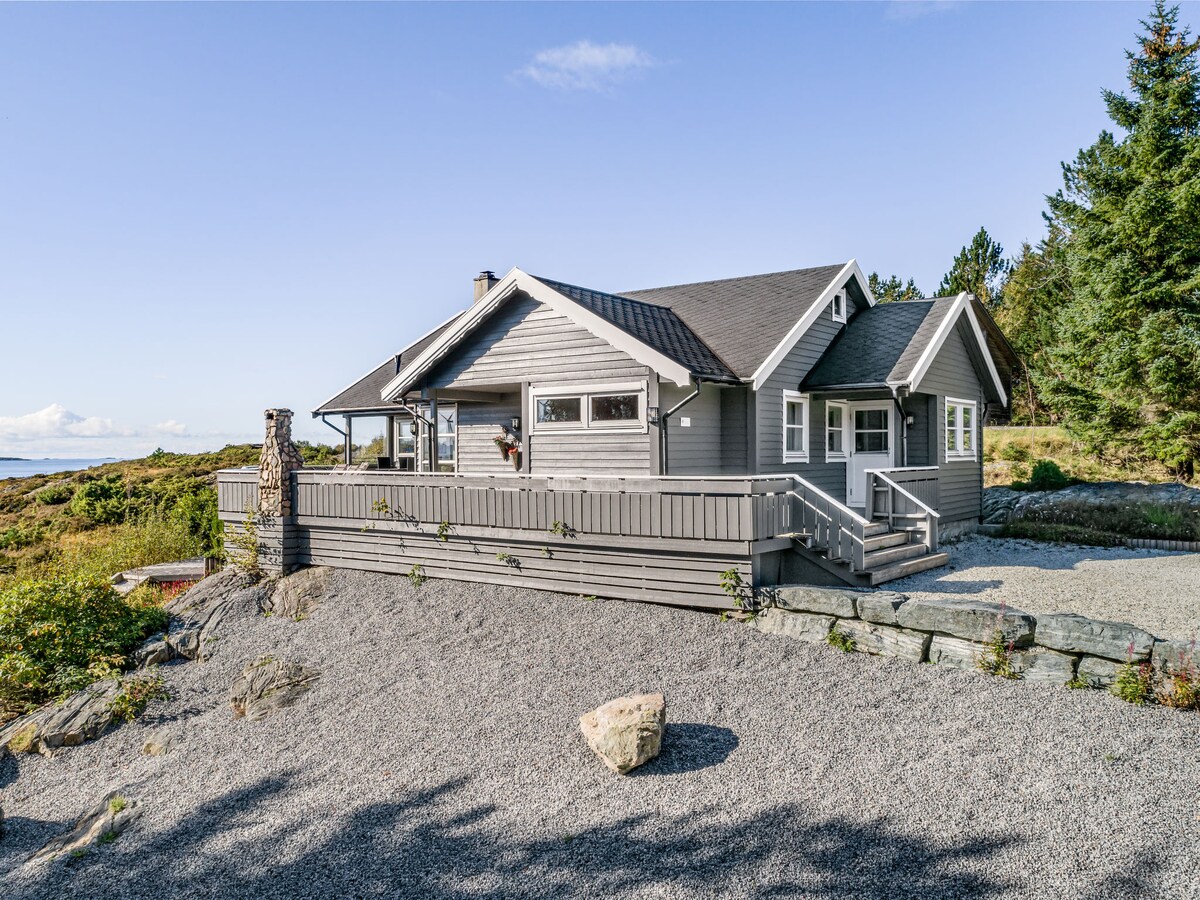 The exterior of the house is presented, showcasing a grey wooden structure with a sloped roof. A spacious terrace is visible, surrounded by a gravel pathway and natural landscaping. Nearby trees provide a sense of privacy, while views of the surrounding area can be seen.