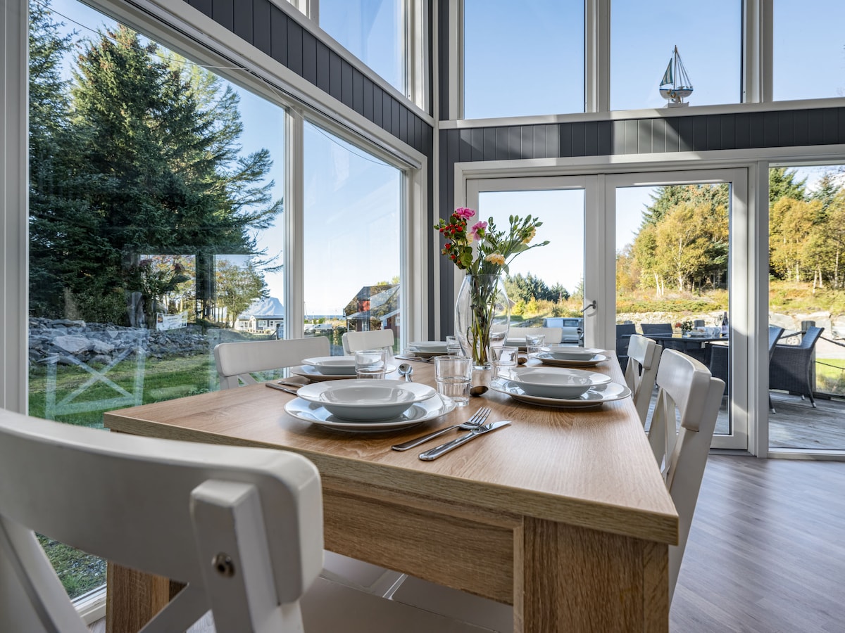 A dining area features a wooden table set with plates and cutlery. Large windows provide ample natural light and views of the outdoor greenery. A vase with flowers adds a touch of elegance to the table setting.