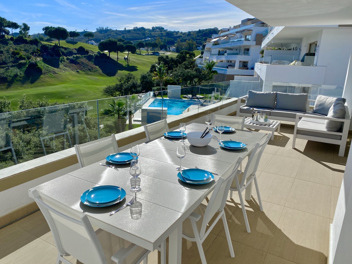 A spacious terrace features a long dining table set with blue plates and glassware. Comfortable seating is provided nearby, with views of the golf course and surrounding greenery. The image captures bright sunlight and a glimmering pool in the distance.