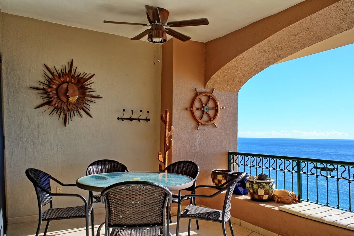A balcony area is displayed with a round glass table and six woven chairs. A ceiling fan hangs above, and decorative wall art complements the space. The ocean can be seen through the archway, offering a serene view of the water and sky.