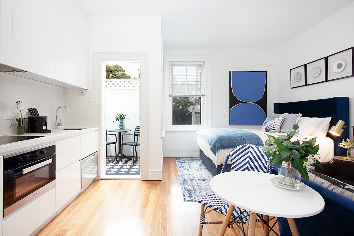 A modern studio features a fully equipped kitchen with sleek cabinetry and stainless-steel appliances. A cozy sleeping area is complemented by a comfortable bed, stylish blue accents, and original wall art. Light enters through a window, enhancing the overall bright and airy ambiance.