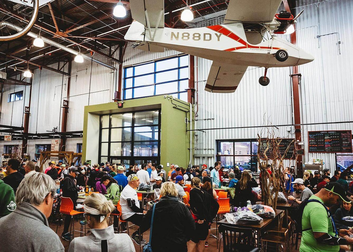 A bustling indoor market is filled with visitors enjoying the atmosphere. An airplane hangs from the ceiling, adding a unique touch to the industrial space, which features large windows allowing natural light to fill the area. Tables are occupied by groups of people dining and socializing.