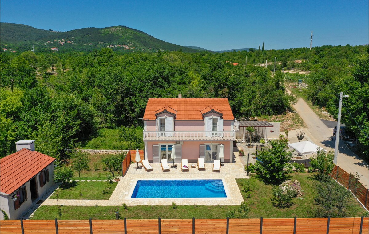 A two-story vacation home is set amidst lush greenery, featuring a private pool surrounded by sun loungers. The building has a red-tiled roof and a spacious outdoor area, accessible through stone pathways, showcasing landscaped gardens and nearby trees.