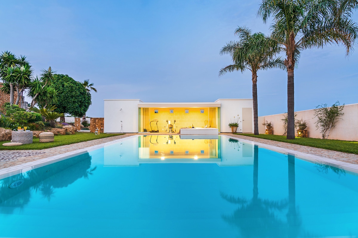 An expansive saltwater infinity pool reflects the surrounding greenery and blue sky. The villa's inviting exterior glows warmly, visible through large glass doors. Palm trees frame the scene, enhancing the serene atmosphere of this peaceful property.