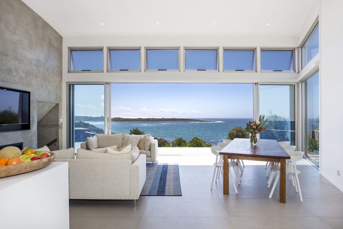 A modern living space is presented with sliding glass doors that open to reveal stunning ocean views. A light-colored sofa is arranged near a wooden dining table, set beside a bowl of fresh fruit. Natural light floods the area, enhancing the spacious ambiance.