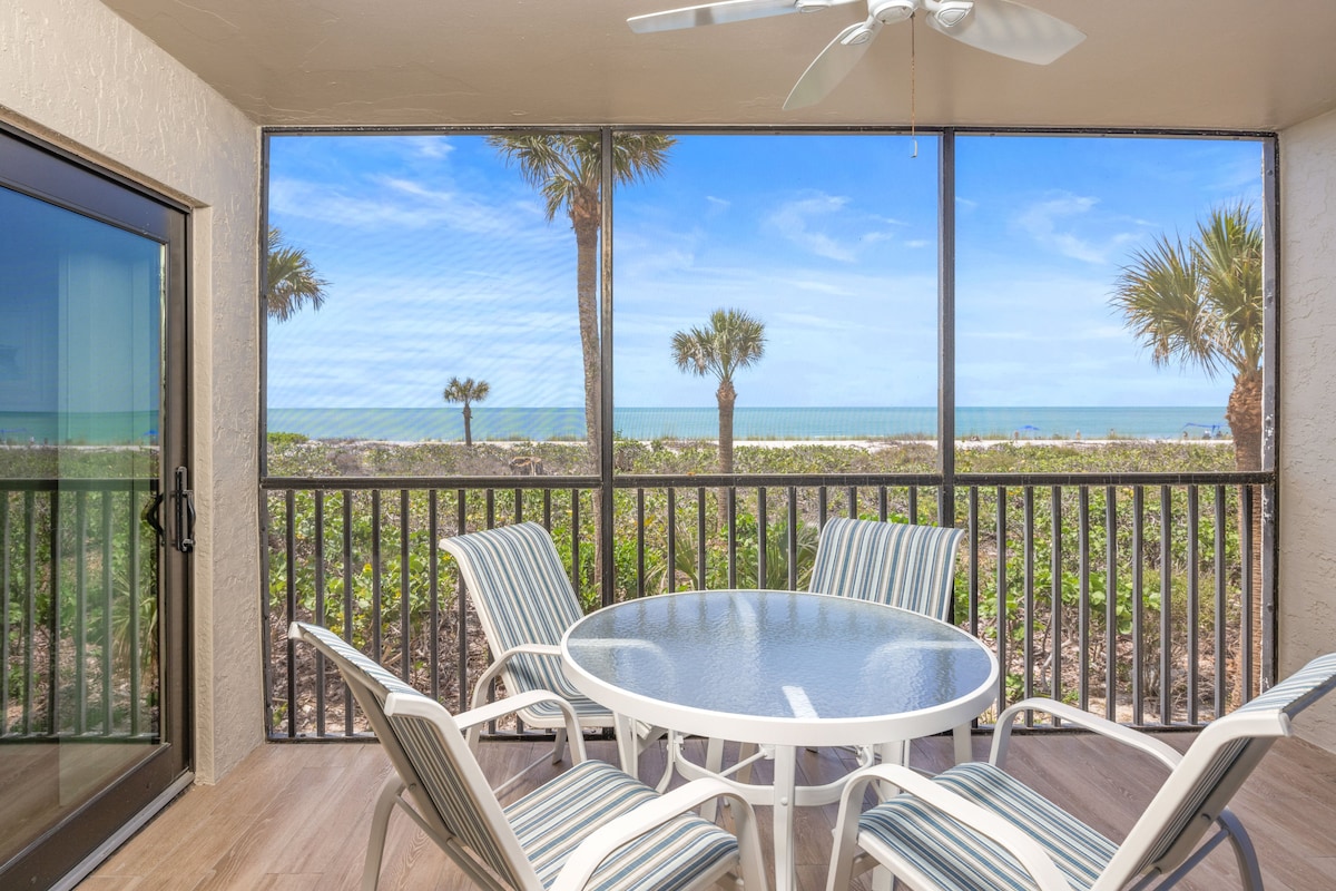 A screened lanai features a round dining table surrounded by six striped chairs. Palms and the calming Gulf of Mexico are visible through the screens, creating a serene outdoor space. Soft natural light enhances the welcoming ambiance.