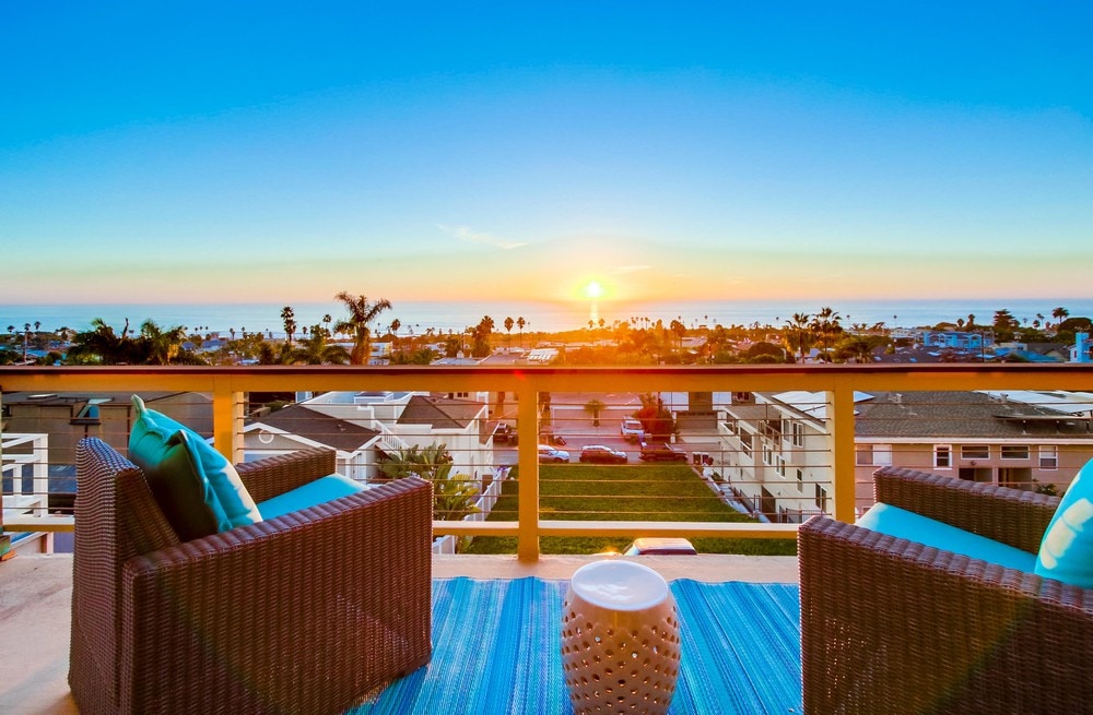 Two outdoor chairs, upholstered in turquoise, are positioned on a deck overlooking a panoramic sunset view of the ocean. A small round table sits between them on a vibrant rug, enhancing the serene ambiance of the space.