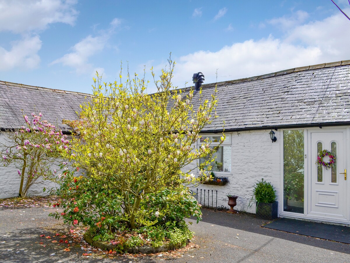 A charming cottage entrance is framed by a circular garden featuring a blossoming shrub. The white stone walls of the cottage contrast with the grey roof under a clear blue sky. A welcoming front door is adorned with a seasonal wreath.