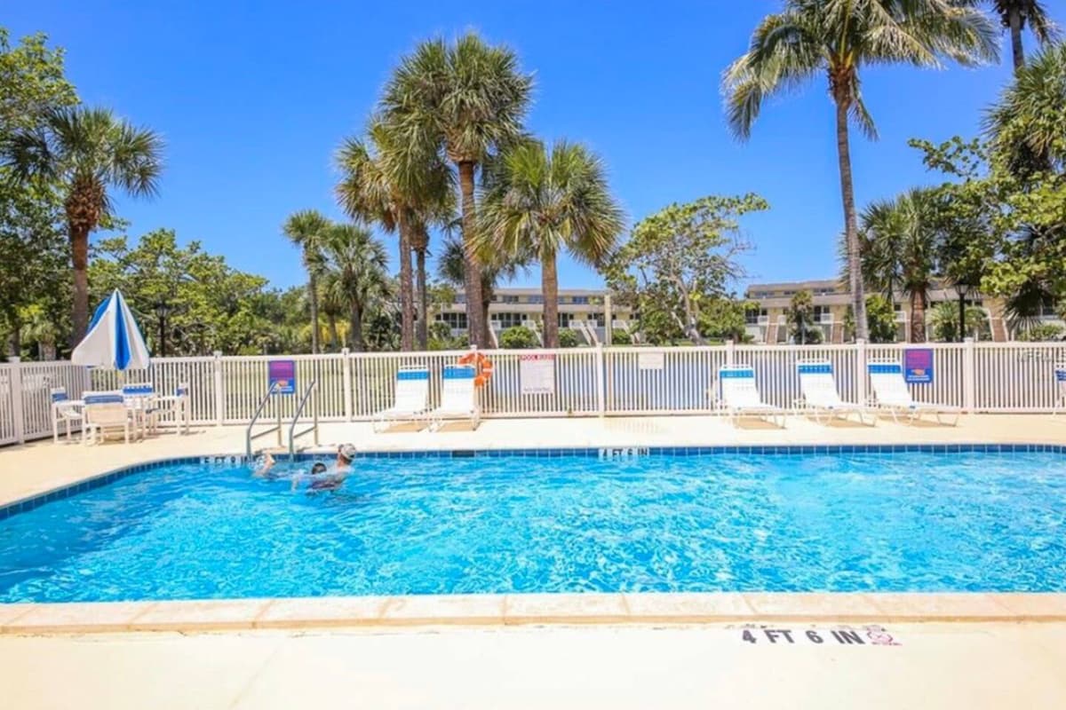 A heated pool area is surrounded by lush palm trees and well-maintained landscaping. Several lounge chairs are arranged around the pool. A swimmer is seen in the water, indicating a family-friendly environment. The clear blue sky enhances the inviting atmosphere of the space.